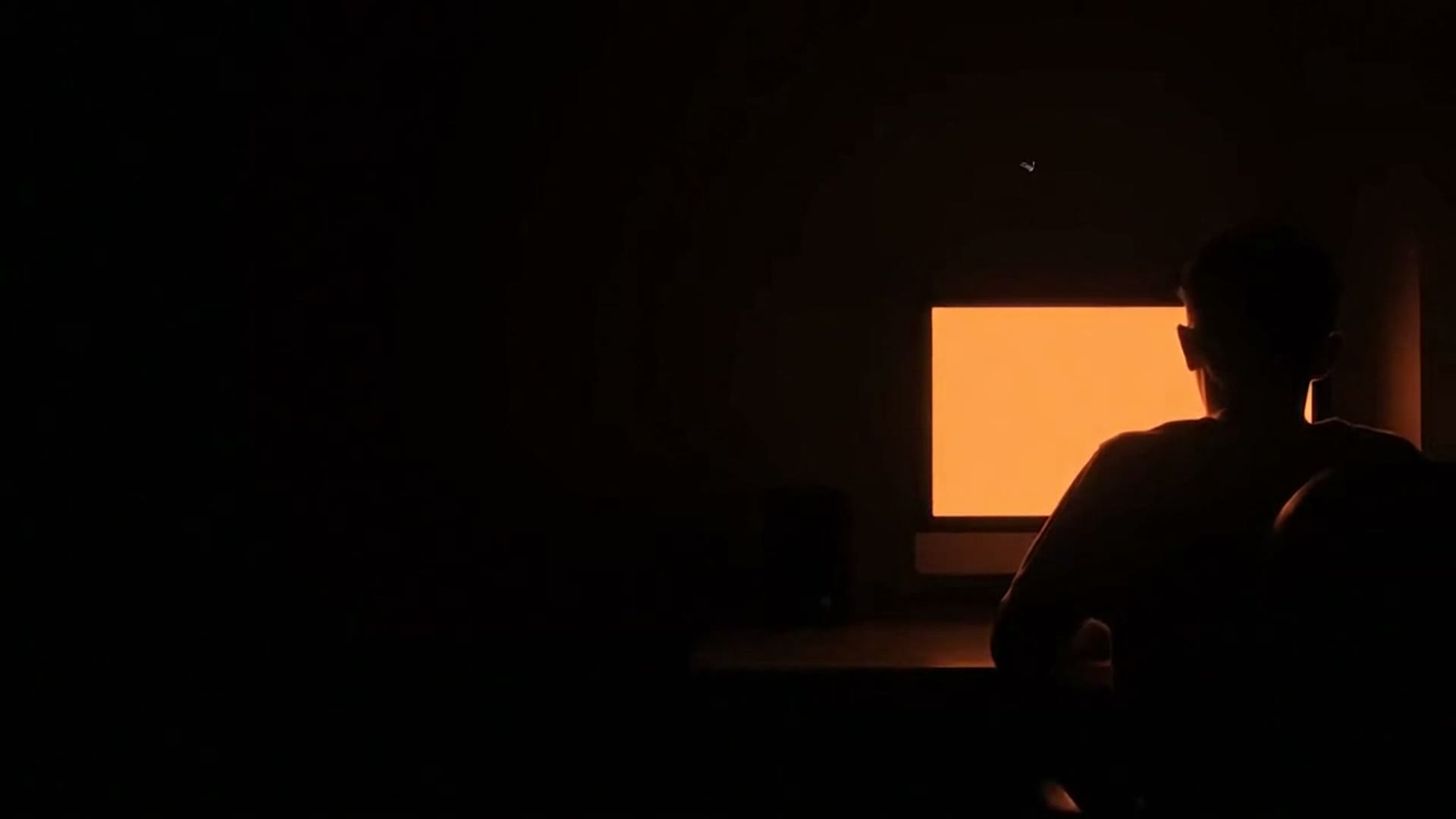 A silhouette at a desk in a dark room, the monitor's ember glow flickering across the walls
